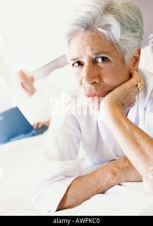 Senior woman on bed with hand under chin, man using laptop in background Stock Photo