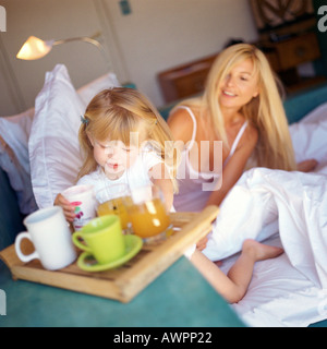 Woman sitting in bed, daughter reaching for glass from tray Stock Photo