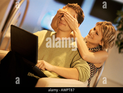 Couple, man using laptop, woman covering man's eyes from behind Stock Photo