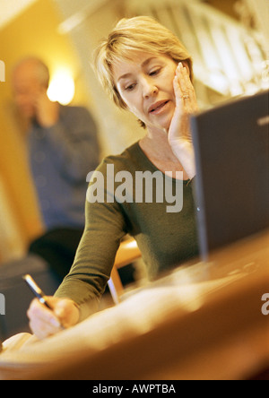 Mature woman using laptop, taking notes, man using cell phone in background Stock Photo