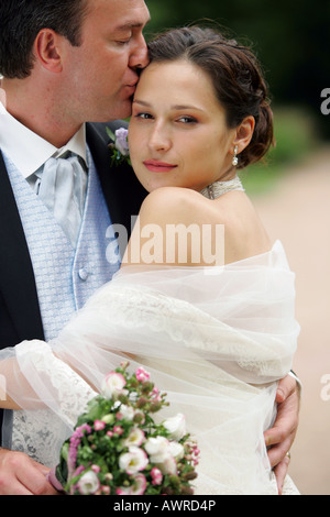 Beautiful smiling bride in white dress with veil on wedding day cuddling her new husband Stock Photo