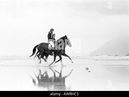 Two people riding horses on beach, side view, b&w.