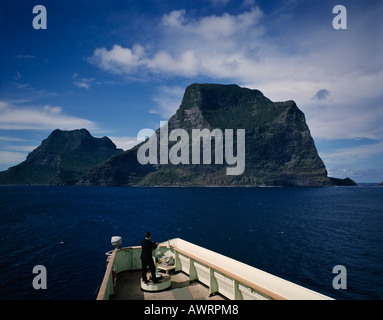 Lone officer on bridge of ship looking across ocean to majestic cliffs of green forested Lord Howe Island in the South Pacific