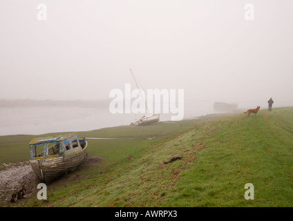 Eerie misty muddy riverbank scene with man walking his Alsatian dog near boats on muddy bank Velator Braunton Pill Devon UK Stock Photo