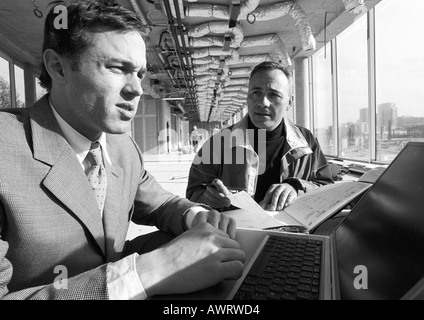 Man looking at agenda, second man using laptop computer, b&w Stock Photo
