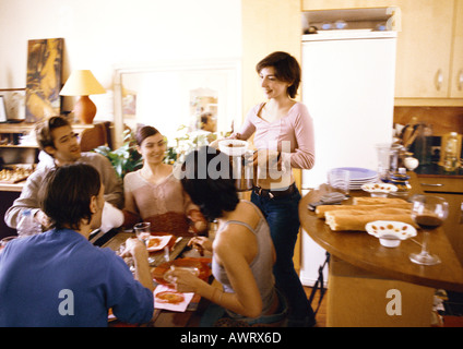Four people sitting at table, woman bringing dish Stock Photo
