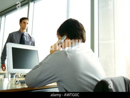 Businessman sitting at computer, using cell phone, second businessman walking in office Stock Photo