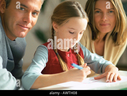 Young girl looking down at table, drawing, between adult man and woman, looking at camera, close-up Stock Photo
