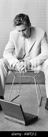 Businessman sitting in chair looking down at laptop on floor, b&w, vertical Stock Photo