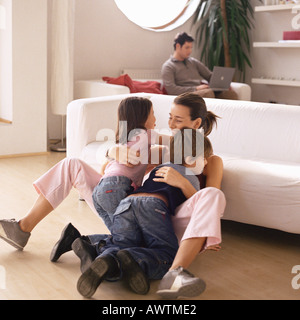 Woman being hugged by two children, man working on laptop in background Stock Photo