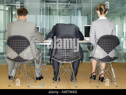 Three business people sitting side by side using computers, one with head down, rear view Stock Photo