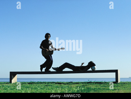 Man playing guitar, teenage girl lying on bench near water Stock Photo
