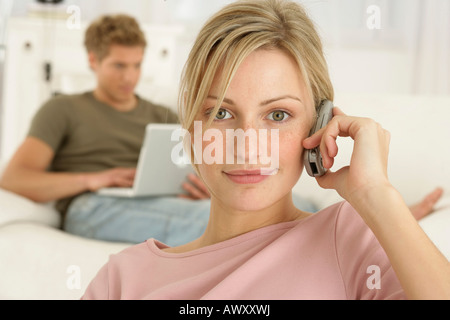 Woman using a phone, man in background sitting on sofa and using laptop Stock Photo