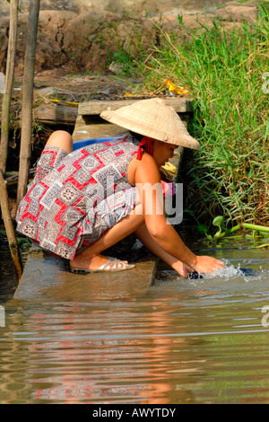 Asia Far East Vietnam , Mekong Delta , pretty young girl in national dress & non bai tho , conical hat , washes clothes in river water Stock Photo