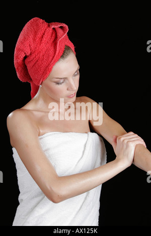 Young woman wearing white towel with red towel wrapped around her head applying body lotion Stock Photo