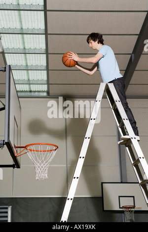A Young Man standing on a ladder preparing to drop a basketball into a basketball hoop Stock Photo