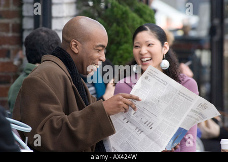 African American man and Asian woman read paper at outdoor cafe Stock Photo