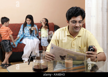 Adult man reading newspaper at dining table with his family sitting in the background Stock Photo