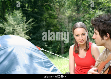 Young couple sitting close to a tent, he is playing the guitar Stock Photo