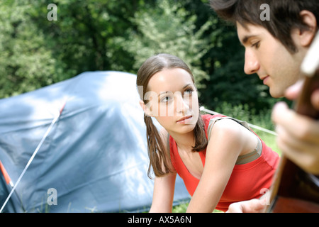 Young couple is sitting close to a tent, he is playing guitar Stock Photo