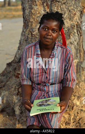 Mbati, Zambia. Woman student in uniform holding a booklet called 'Zambia's Pride', sitting at the foot of a tree. Stock Photo