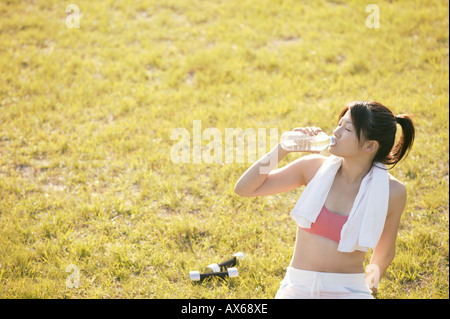 A young woman drinks water as she sits amidst grassland after her workout Stock Photo