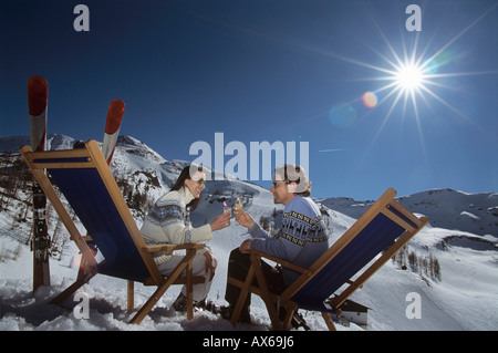 Couple sitting on deck chairs in snow, holding glasses, side view Stock Photo