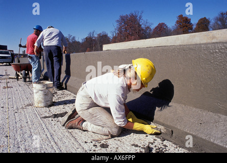 Female Construction Worker Smoothing Wet Concrete on New Interstate Highway Barrier while Male Workers Examine Barrier Stock Photo