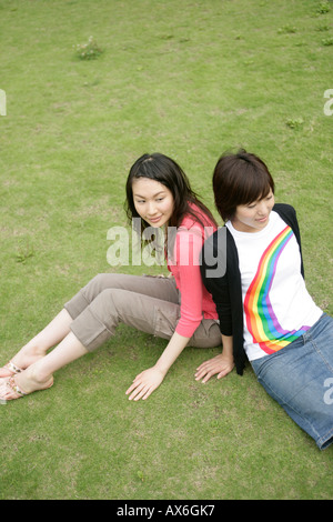 Two young women rest on each others back as they sit on the grassland Stock Photo