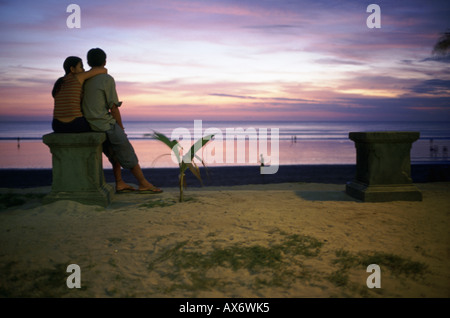 A young couple sits near the beach during sunset at Kuta Beach Bali Indonesia Stock Photo