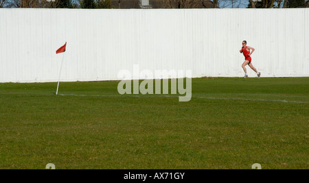 Man in red shorts and top jogging on a cricket club pitch past white fence towards red corner flag, Cheam, south London, Surrey Stock Photo