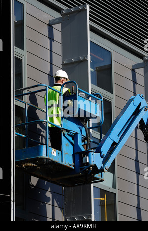 A construction worker in a lift wearing a hard hat and safety vest is working on the exterior of a building using hydraulic lift or cherry picker Stock Photo