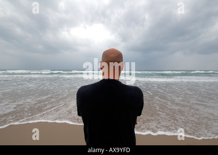 a bald man, viewed from behind, standing on a sandy beach and looking out at an ocean with rolling waves under a dark, overcast sky Stock Photo