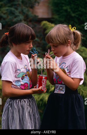 Two young twin sisters sip Coke from their own curly straws in an English garden, UK Stock Photo