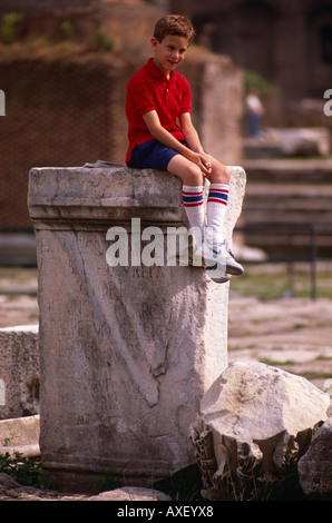 Boy tourist sits on top of an ancient statue pedestal in the ruins of the Roman Forum in central Rome, Italy