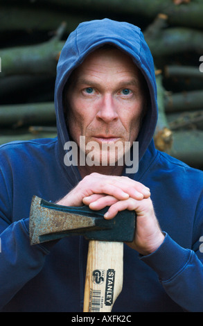 man in his forties wearing a blue hooded sweatshirt is posing with a woodcutting axe in front of a woodpile outside Stock Photo
