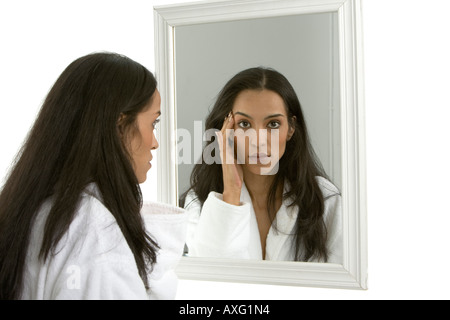Attractive young woman with long black hair looking at herself in the mirror wearing a white fluffy bathrobe Stock Photo