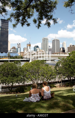 Two women sitting on grass bank with Central Business District beyond in summer Brisbane Queensland QLD Australia Stock Photo