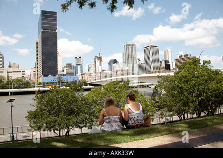Two women sitting on grass bank with Central Business District beyond in summer Brisbane Queensland QLD Australia Stock Photo
