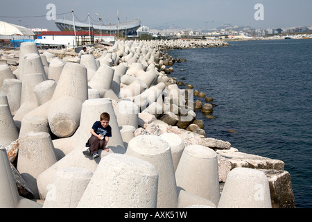 Young boy climbing on sea wall defences by Mikrolimano with Peace and Friendship stadium beyond in Piraeus or Pireas Athens Gree Stock Photo