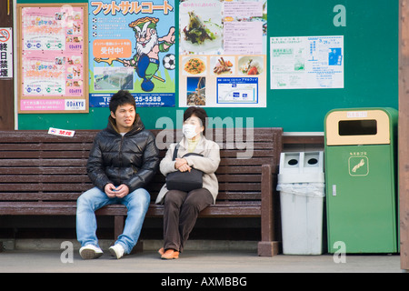 Japanese man and woman sitting on bench waiting for train with one wearing mask over her face for personal hygiene Stock Photo