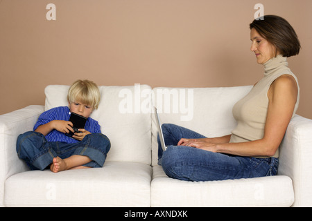 Mother and son (4-5 Years) sitting on a sofa, woman using a laptop, boy playing hand held computer game Stock Photo