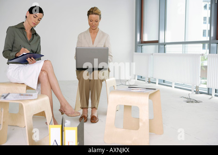 Two business women sitting on stools, one working on laptop, the other is writing something down Stock Photo