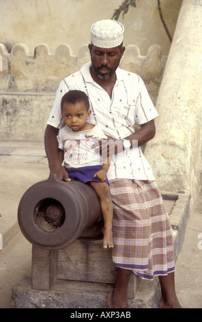Father holding his young son whilst he sits on an ancient cannon outside the Omani Fort on Lamu Island Kenya East Africa Stock Photo