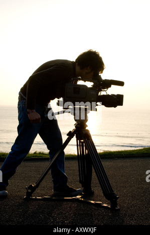 Silhouette man student Aberystwyth University TV and Film studies department using video camera on  tripod at seaside, Wales UK Stock Photo