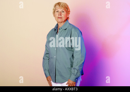 Woman in her 60s, sixty something years old stands against a colorfully lit wall. Stock Photo