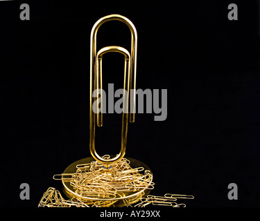 A pile of small gold paperclips lay on the stand of a large gold paperclip.  They are set against a black background. Stock Photo