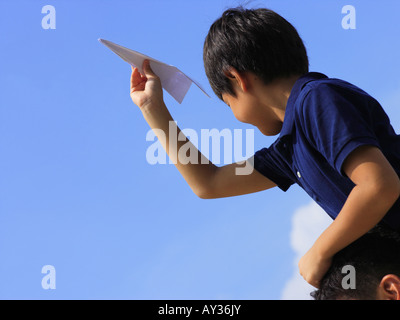Low angle view of a boy sitting on his father's shoulders flying a paper airplane Stock Photo