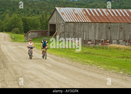 Couple riding mountain bikes on a rural dirt road passing a weathered barn in Warren Vermont on an overcast summer day Stock Photo