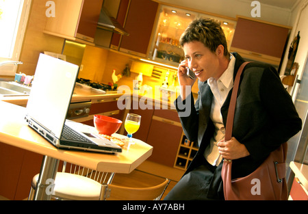 Businesswoman multitasking on phone with computer laptop in the kitchen in the morning rush after breakfast Stock Photo